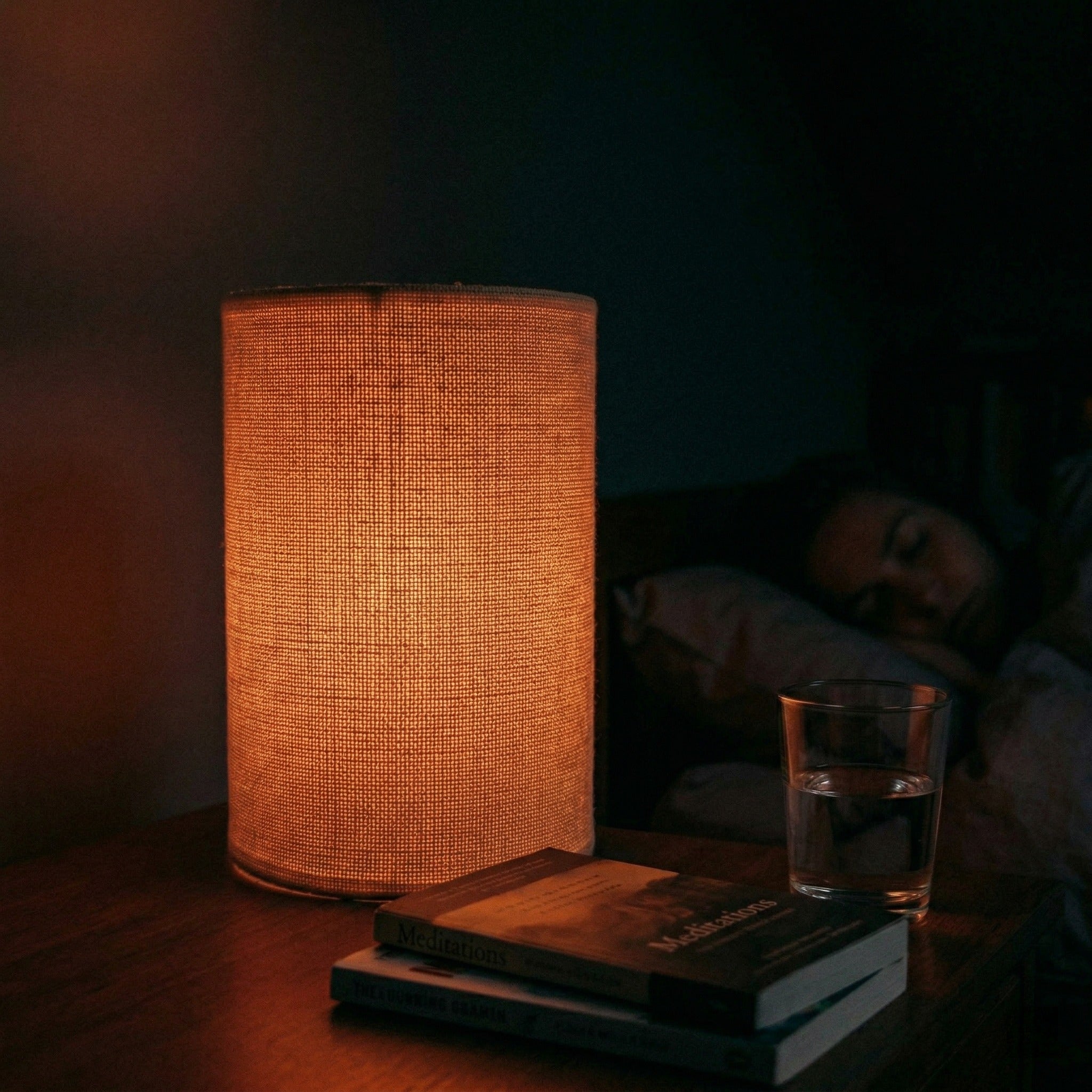 Lamp on a table with books and a glass of water, person lying in bed in the background