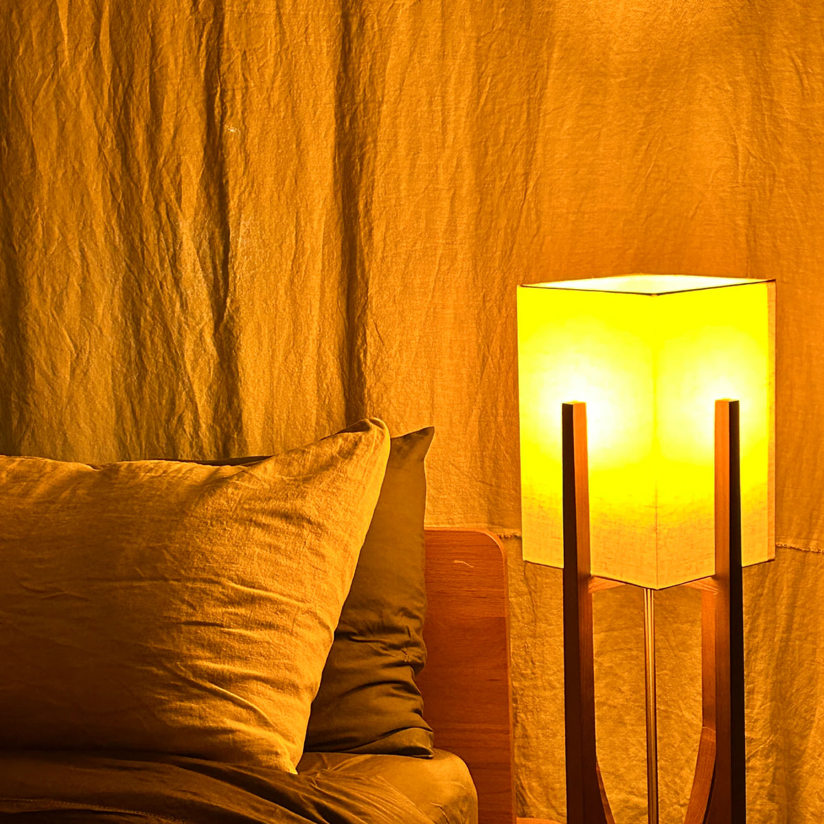 Bedside lamp glowing warm amber in a darkened bedroom, lighting linen pillows and a draped curtain behind the bed