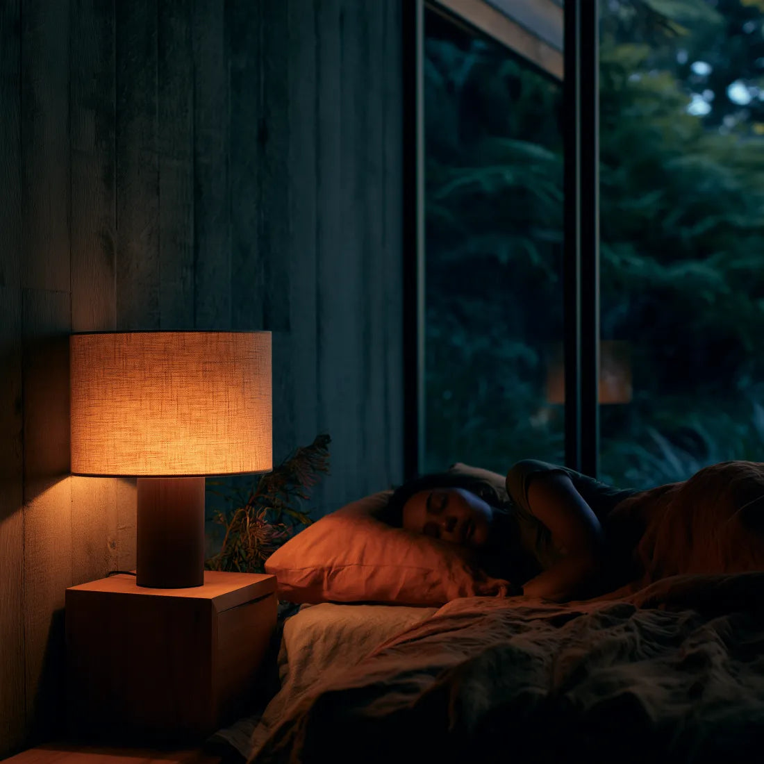 A warm glowing table lamp on a wooden bedside table next to a bed where a person is sleeping, in a dark, cozy bedroom setting.