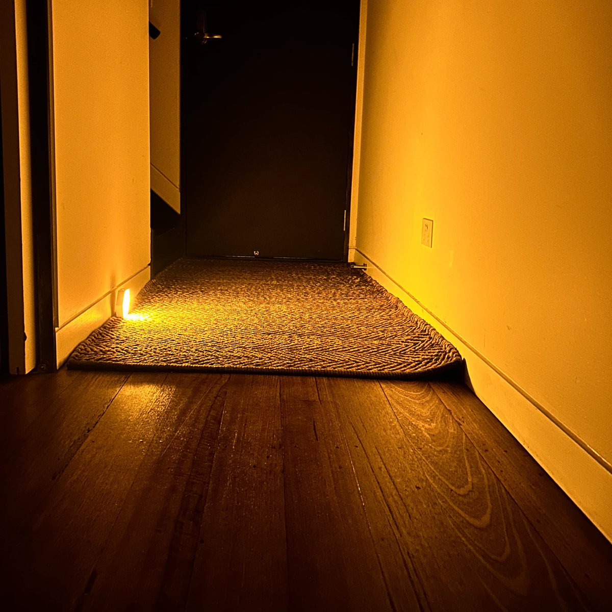Motion-activated amber night light glowing at floor level in a hallway doorway, lighting a jute rug and timber floorboards at night