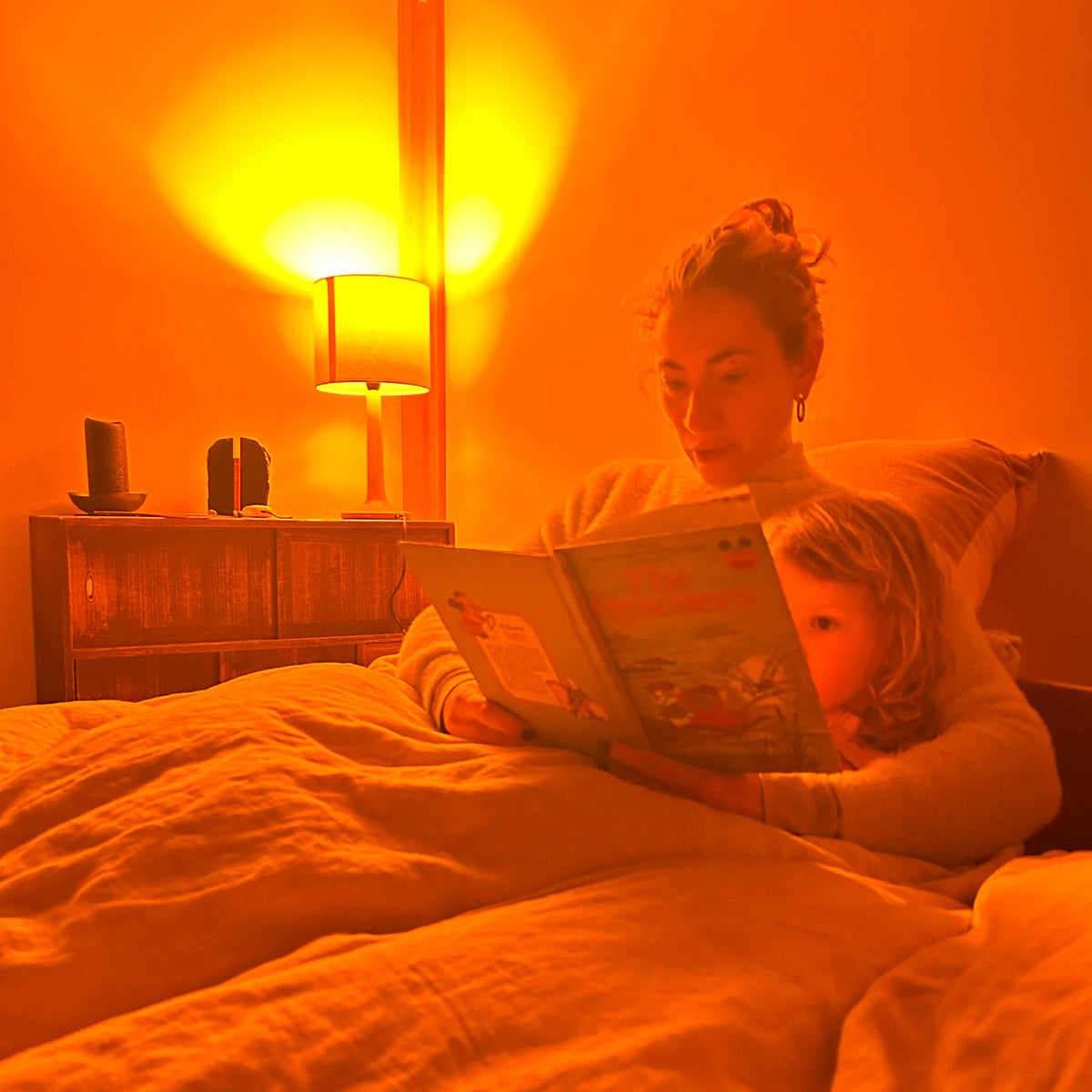 A woman and a young child reading a picture book together in bed at bedtime, lit only by the warm amber glow of a bedside lamp