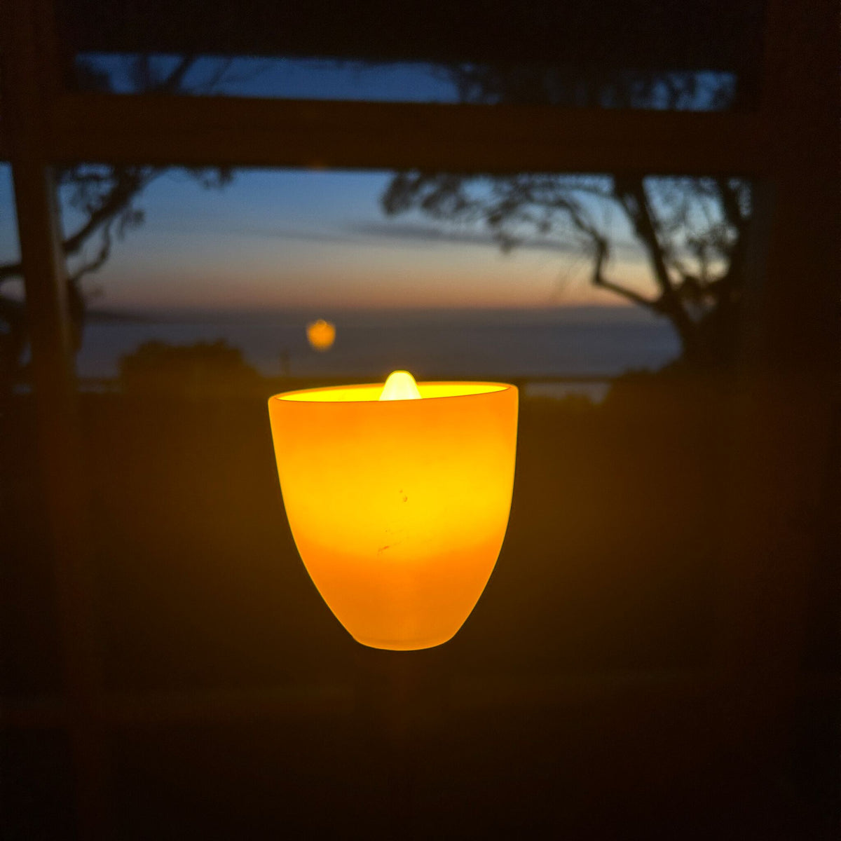 Lamp with an E14 amber bulb glowing in front of a sunset over the water, seen through a window at dusk