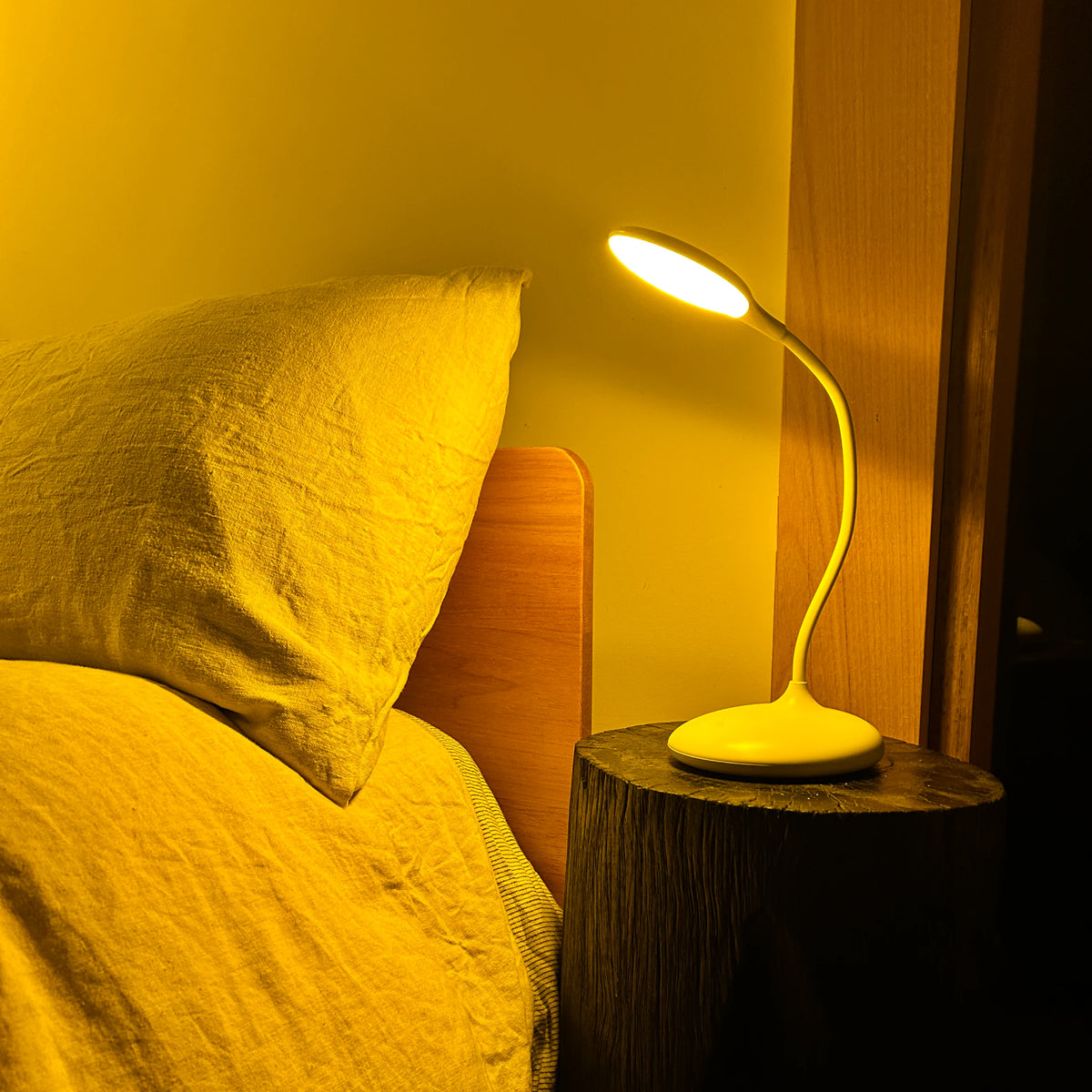 Blue blocking amber sleep lamp with a flexible gooseneck arm, glowing warm amber on a timber stump bedside table beside a linen-dressed bed at night
