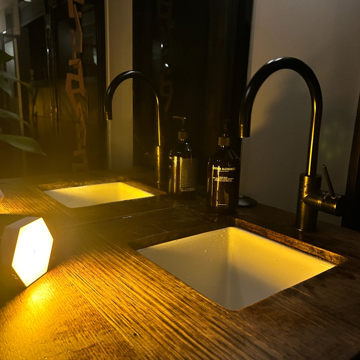 Hexagonal motion-activated amber night light on a timber bathroom vanity, lighting twin basins and black gooseneck tapware with a warm amber glow at night