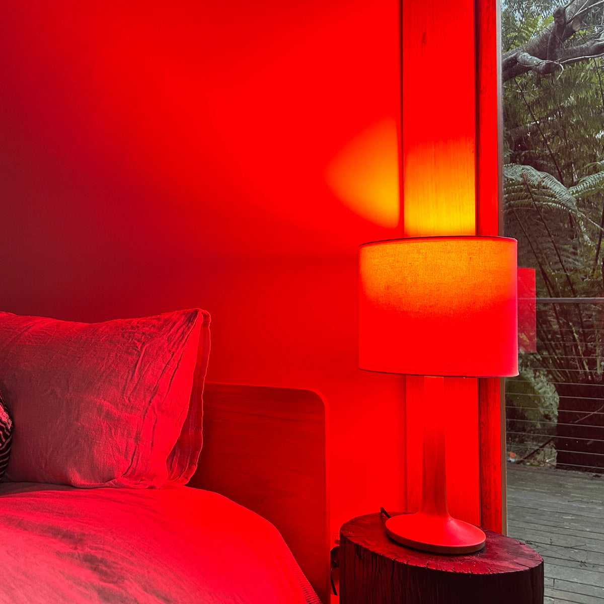 Bedside table lamp fitted with a red night light bulb, filling a bedroom with deep red light across linen bedding and a timber headboard, with tree ferns visible through the window at night