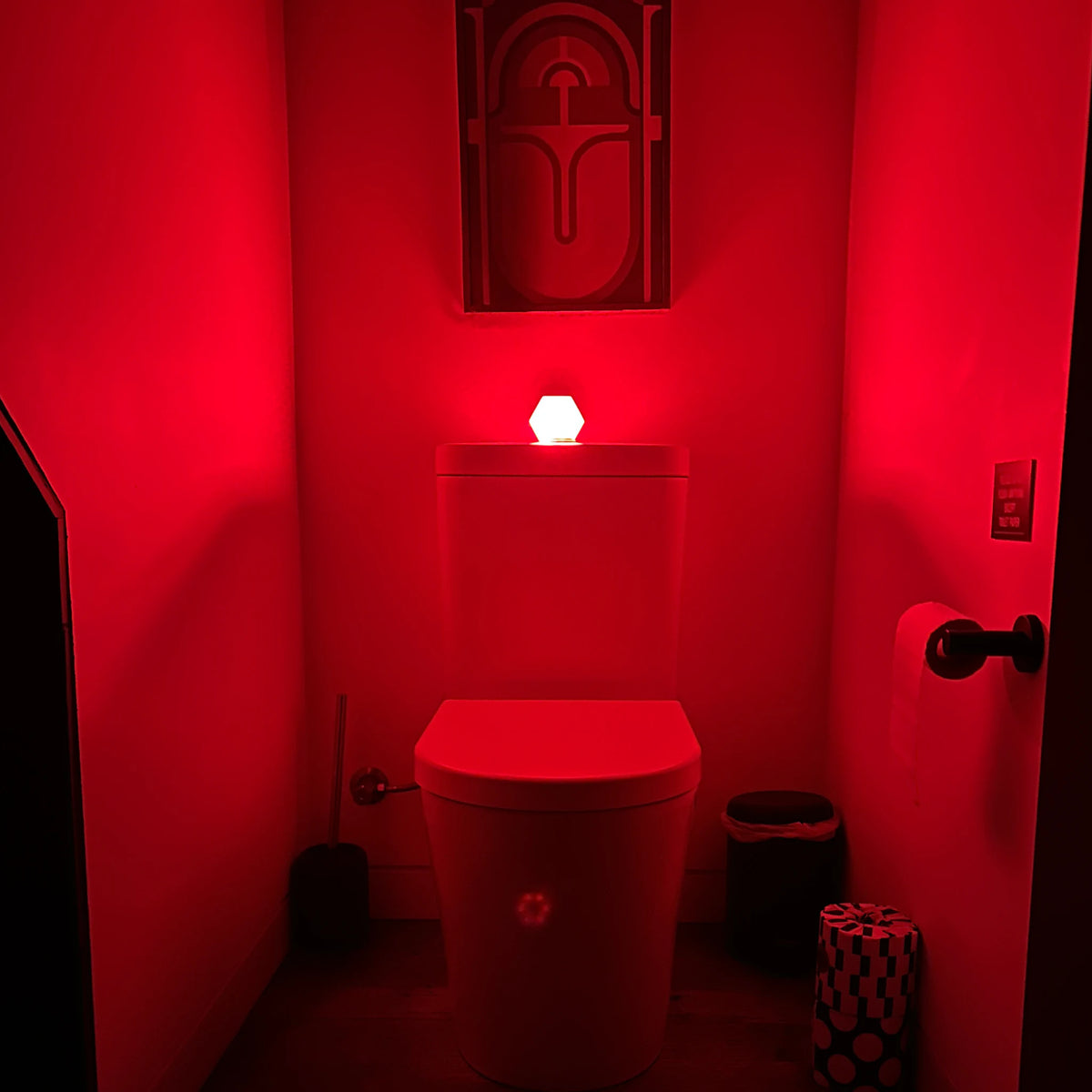 Hexagonal motion-activated red night light sitting on a toilet cistern, lighting a small bathroom entirely in red for a night-time visit