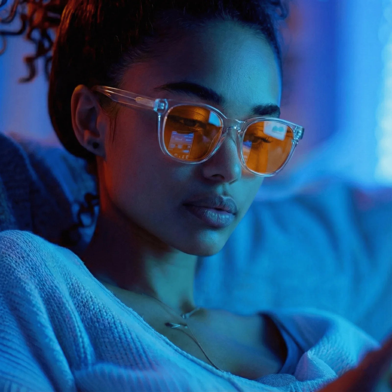A woman wearing clear-framed sunglasses with orange-tinted lenses, shown in a close-up lifestyle shot under blue ambient lighting.
